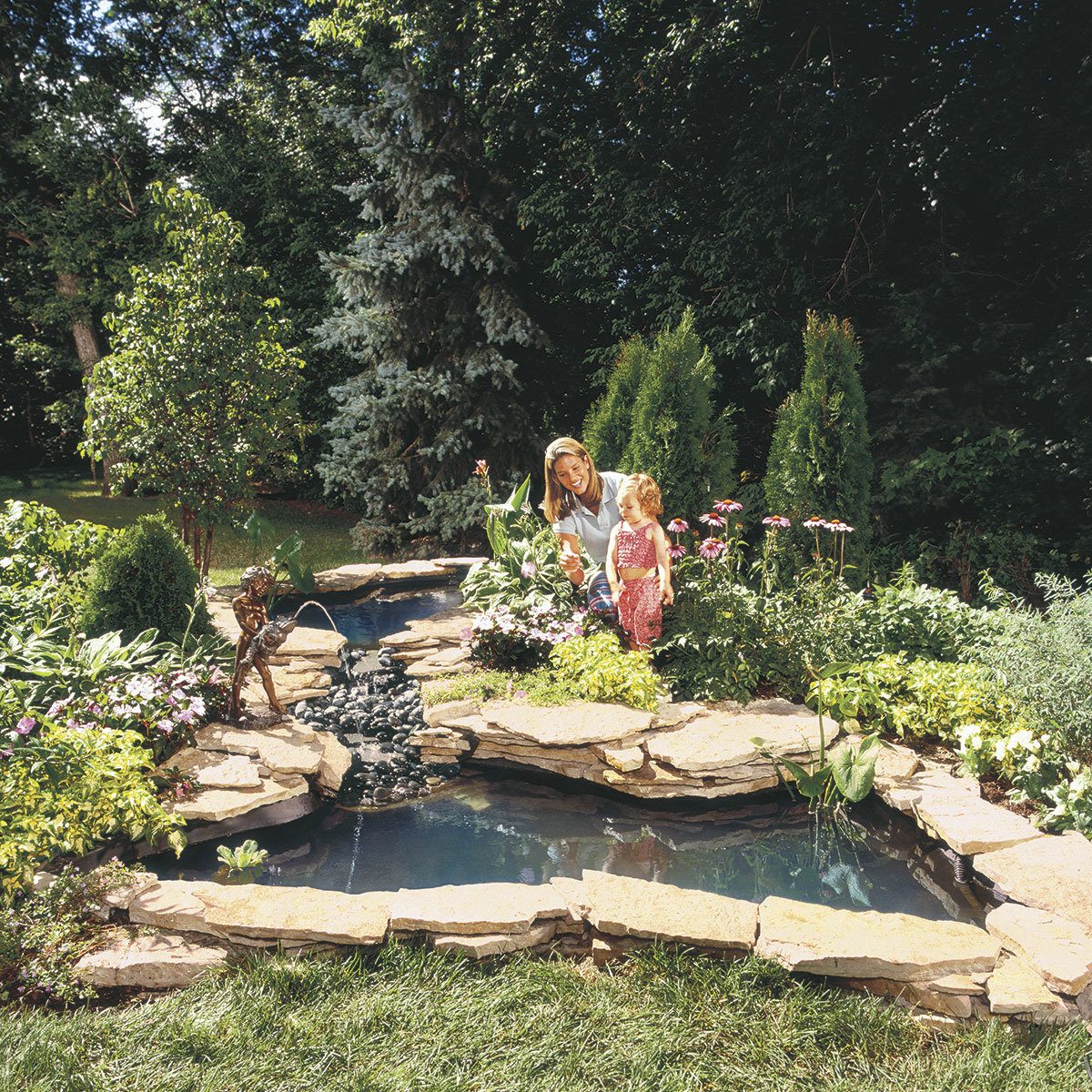 A woman and a child tend to a garden around a pond with flowers, stones, and greenery under a sunny sky in a tranquil outdoor setting.