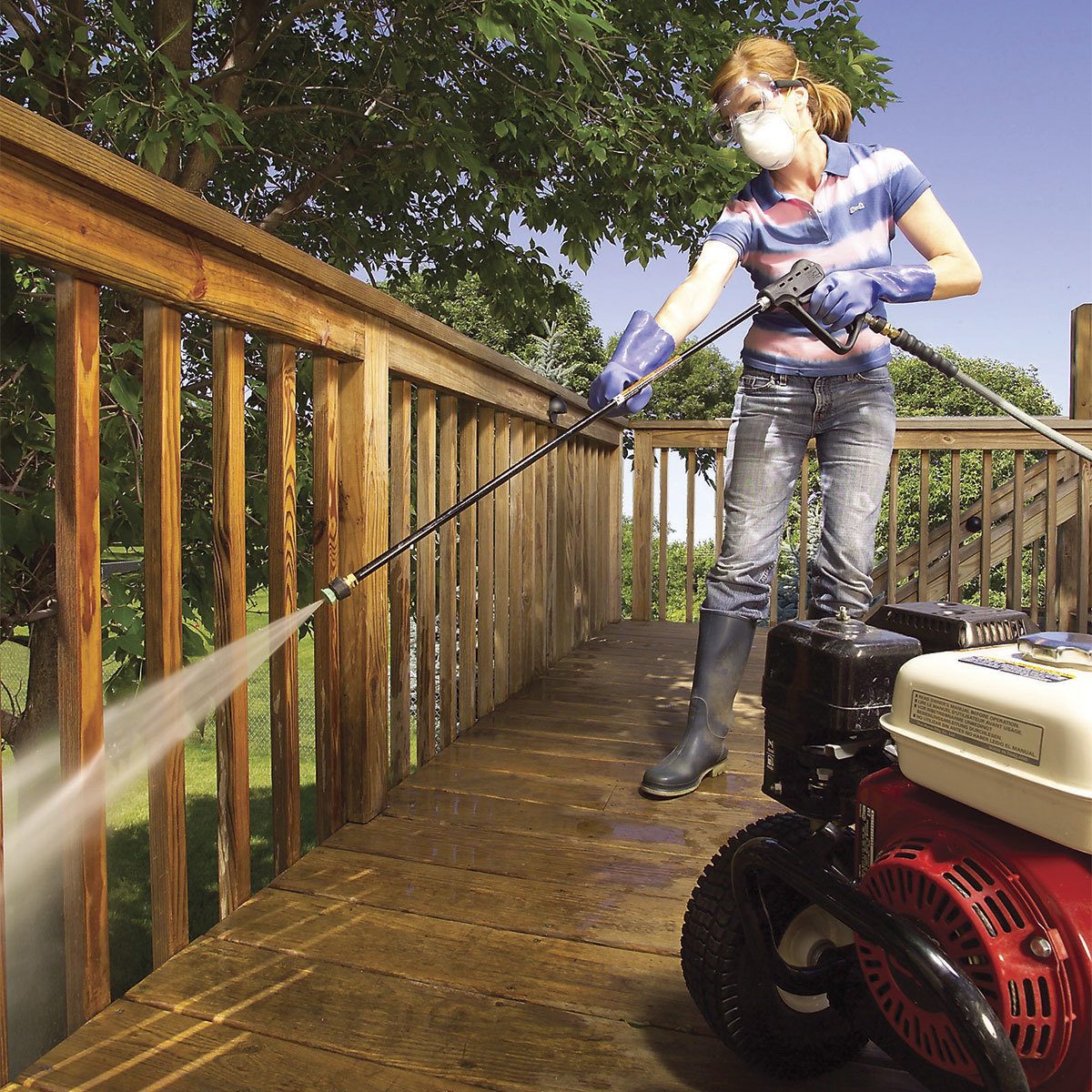 A woman uses a pressure washer on a wooden deck, cleaning it while surrounded by trees and bright blue skies. She wears protective gear.