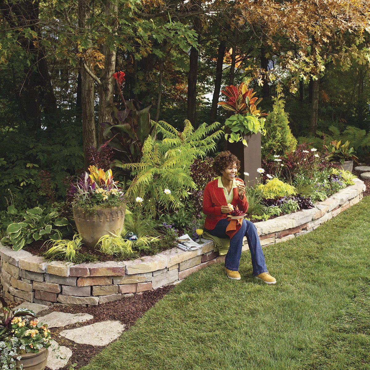 A woman sits on a stone wall, enjoying flowers and nature in a vibrant garden filled with various plants, surrounded by trees and grass.