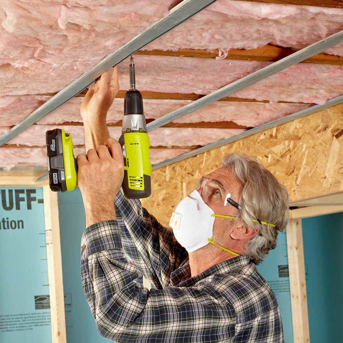 A man uses a cordless drill to secure metal framing to a ceiling filled with pink insulation, working in a partially constructed room.