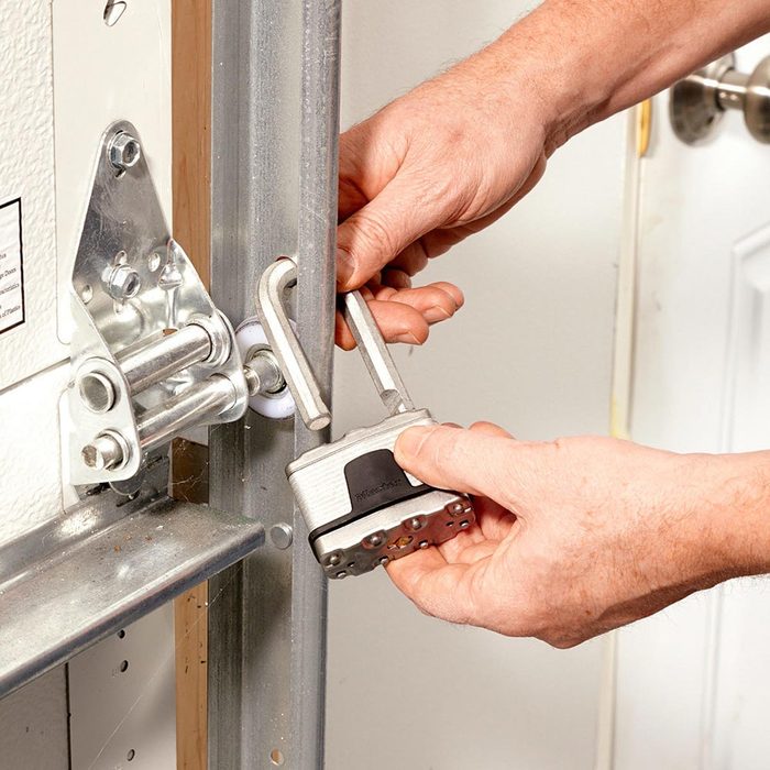 A person closes a padlock on a door's metal frame, securing it in place, within a room featuring a door and a metallic track mechanism.