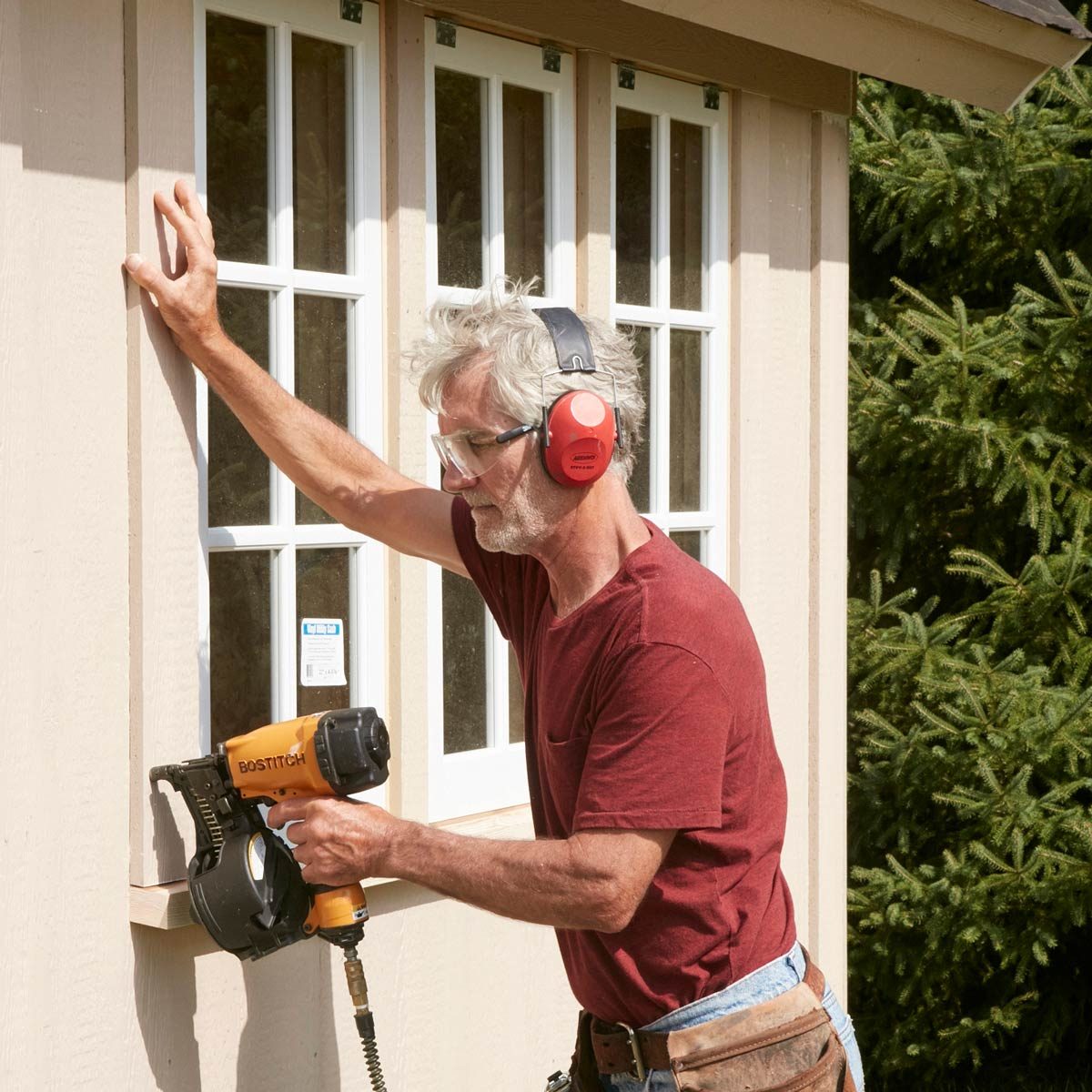 A man uses a nail gun to attach a trim piece to a building wall, wearing safety glasses and ear protection, with trees in the background.