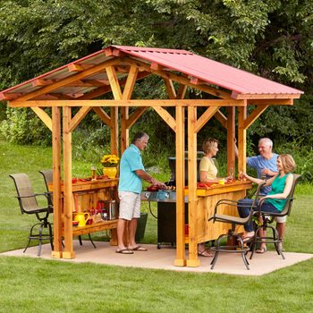 A wooden pavilion with a red roof hosts four people socializing, while one cooks on a grill. They're surrounded by a grassy outdoor setting.