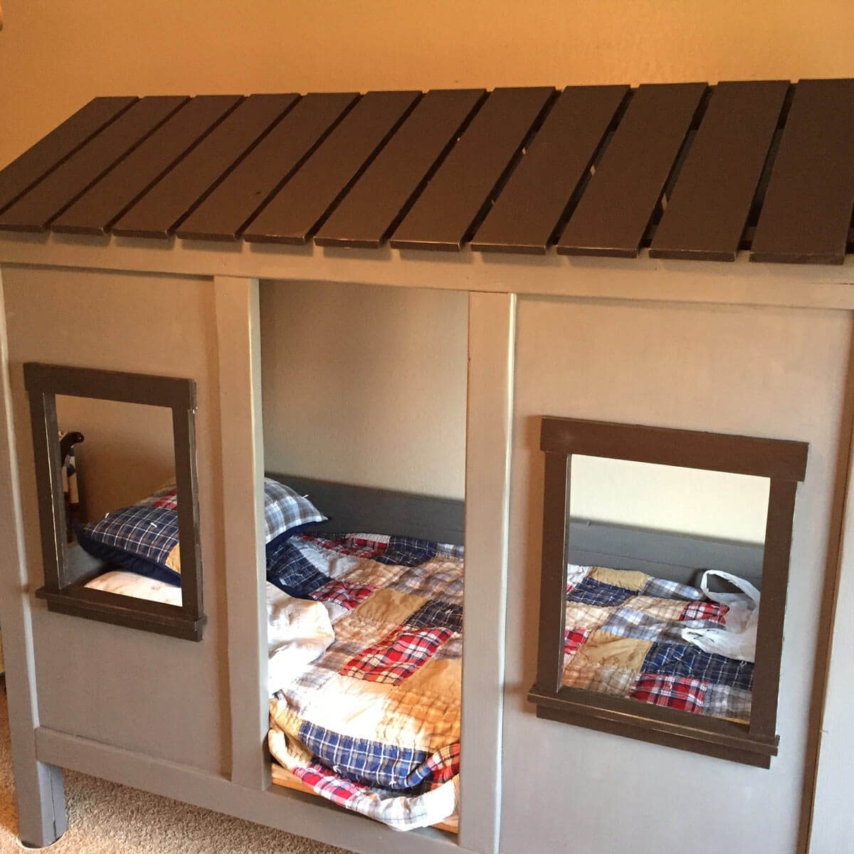 A wooden playhouse bed with slanted roof, featuring two open windows and a colorful quilted blanket on the mattress, set against a plain wall.