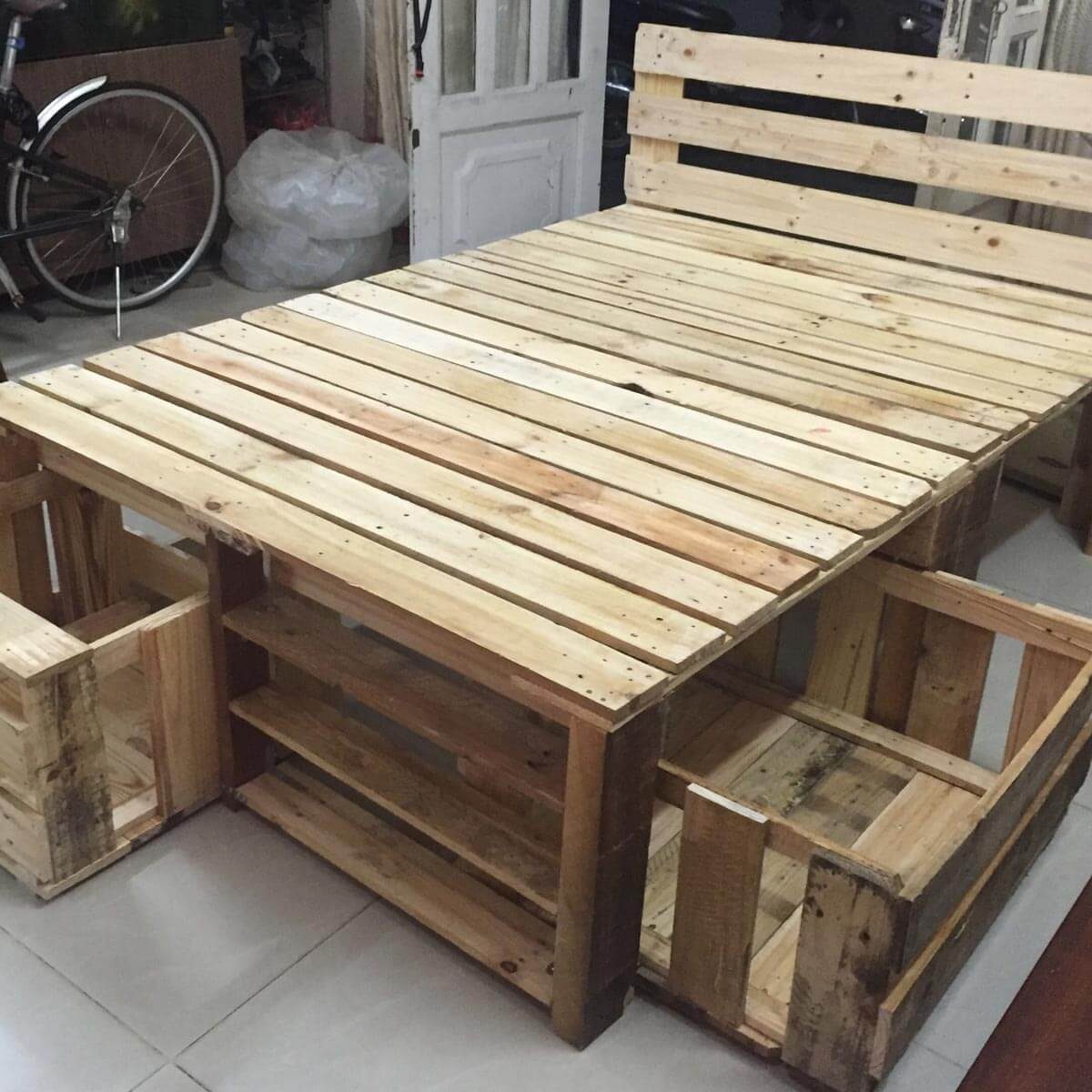 A wooden pallet table with attached seating sections rests on a tiled floor, surrounded by a bicycle and a bag in a cozy indoor setting.