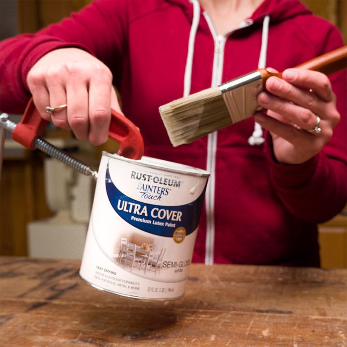 A person grips a paint can tightly while holding a paintbrush, preparing to paint in a workshop-like environment with wooden surfaces.