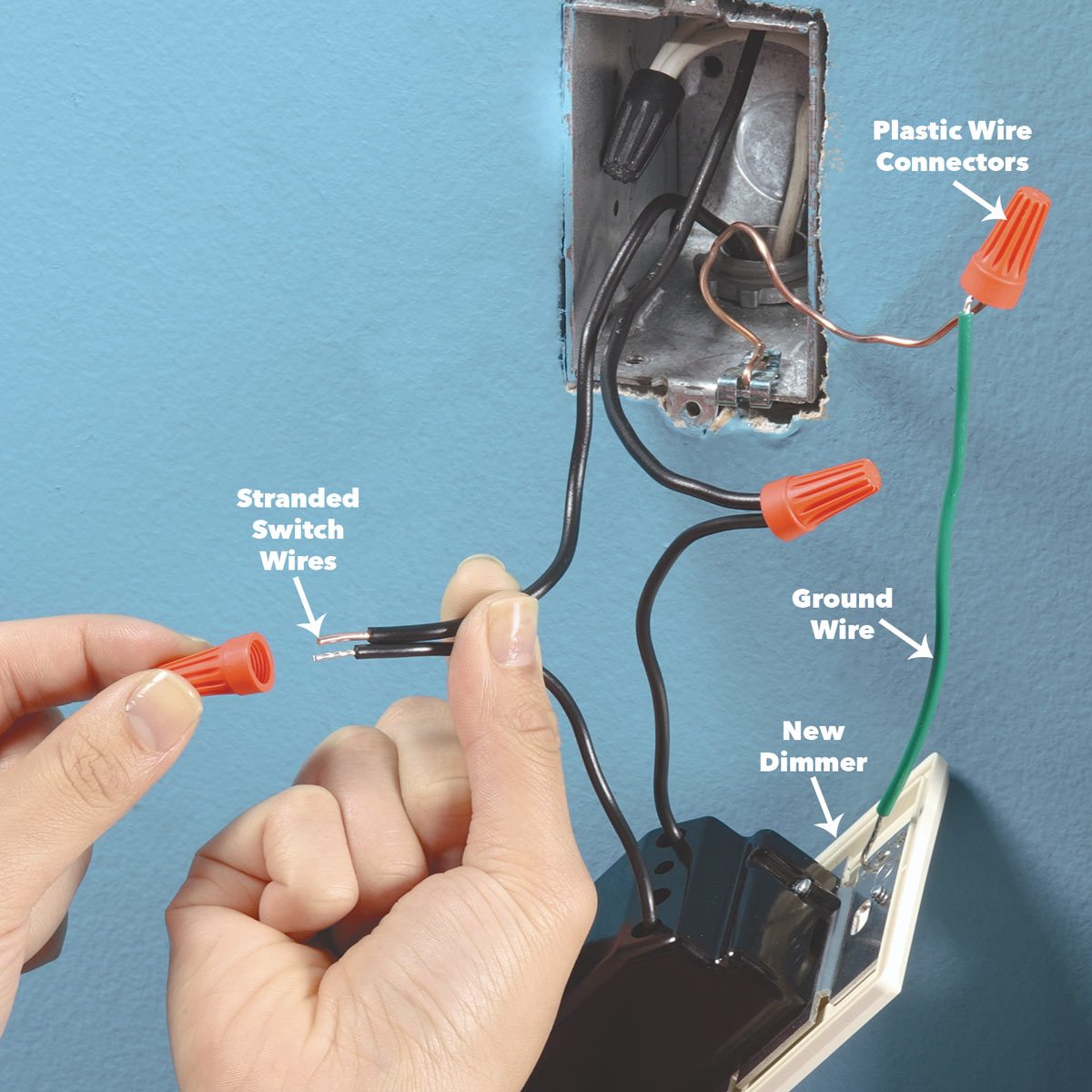 A hand connects a red plastic wire connector to stranded switch wires near a newly installed dimmer switch on a blue wall.
