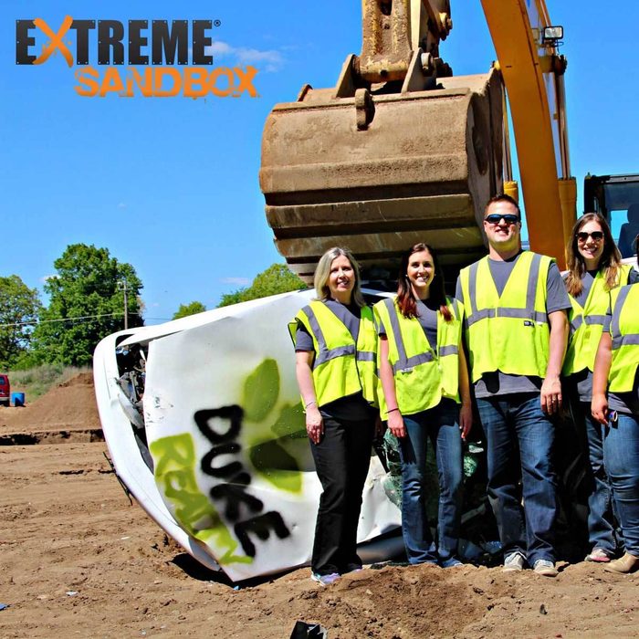 A group of five people in yellow vests stands near a toppled vehicle, while a large excavator looms above in a dirt-filled outdoor area.