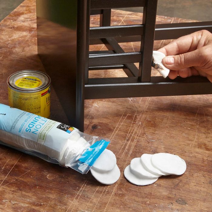 A hand is cleaning the leg of a black wooden chair with a cotton pad, surrounded by a can and a package of cotton rounds on a wooden surface.