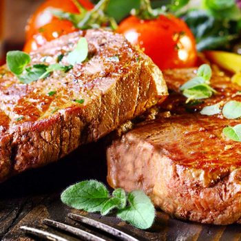 Two grilled steaks garnished with herbs sit on a wooden surface, accompanied by cherry tomatoes and a colorful vegetable salad, with a glass beverage nearby.