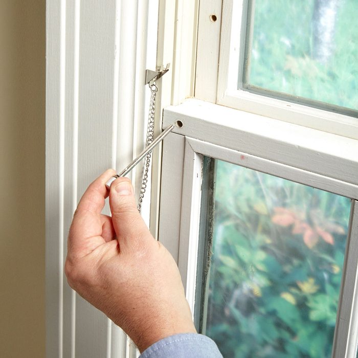 A hand holds a screwdriver, securing a window latch into a hole on a white window frame, with foliage visible through the glass.