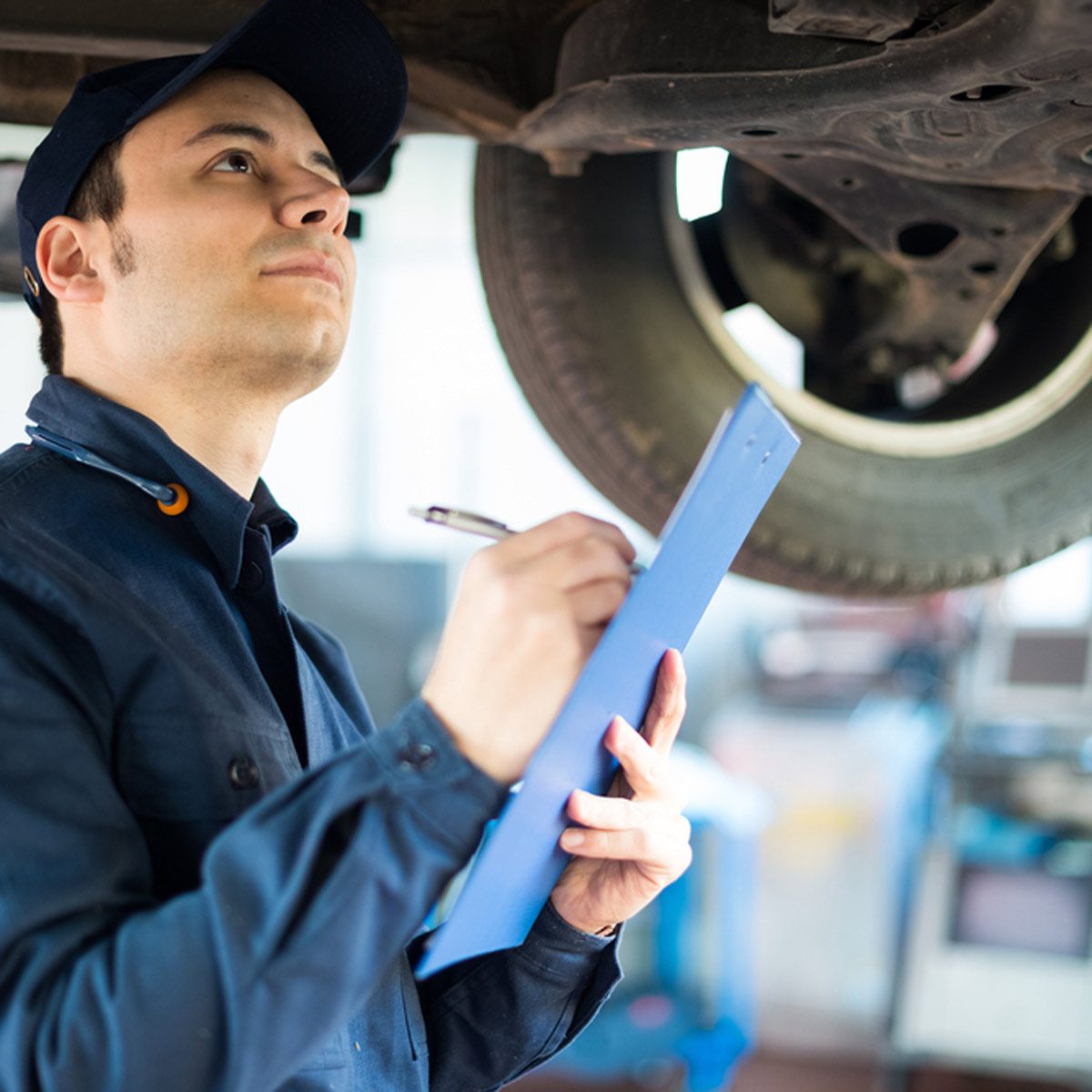 A mechanic inspects a vehicle's undercarriage, jotting notes on a clipboard in a well-lit garage with tools and equipment visible in the background.
