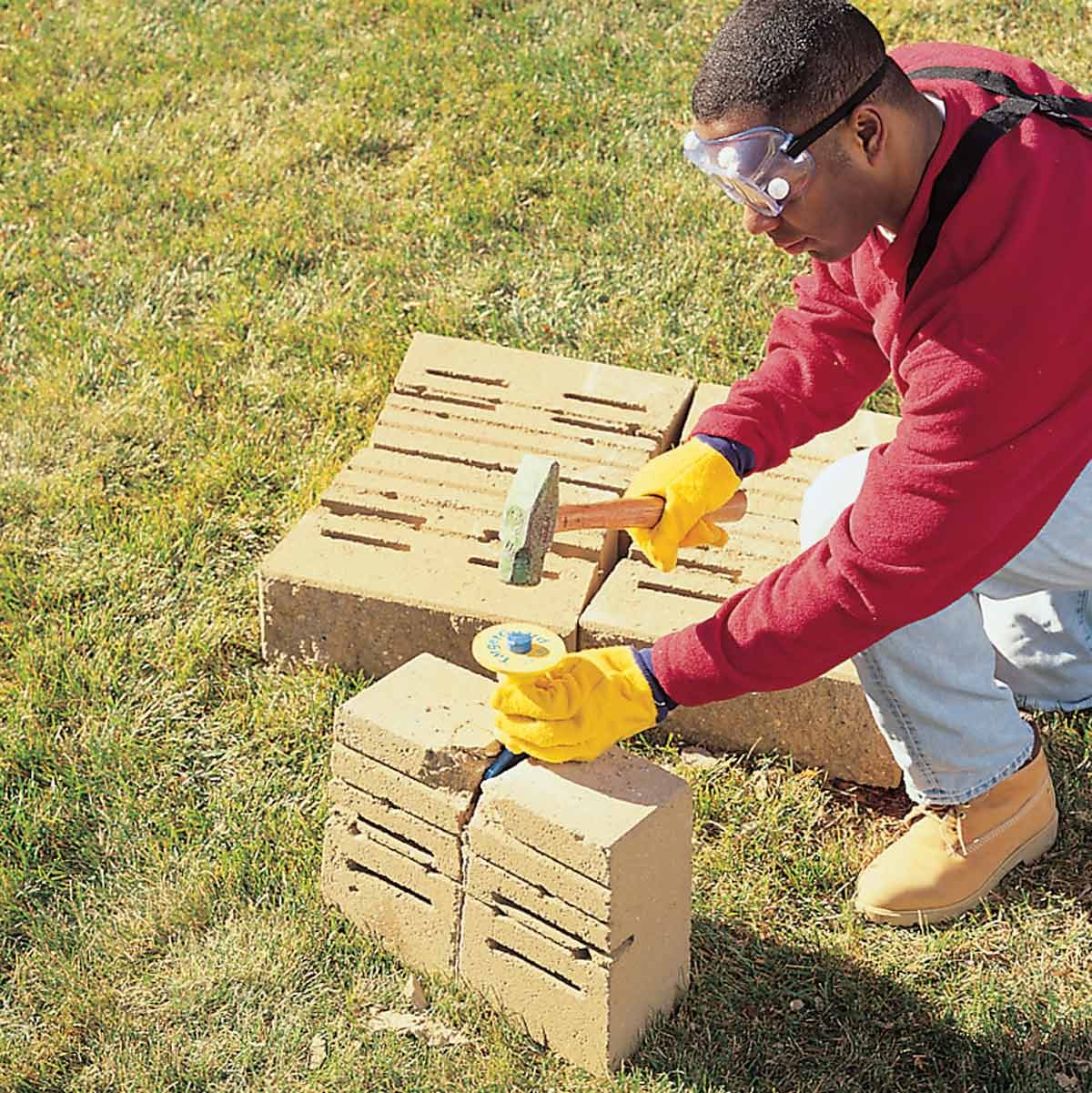 A man wearing gloves and goggles is chiseling a concrete brick on a grassy surface, using a hammer to break apart the material.