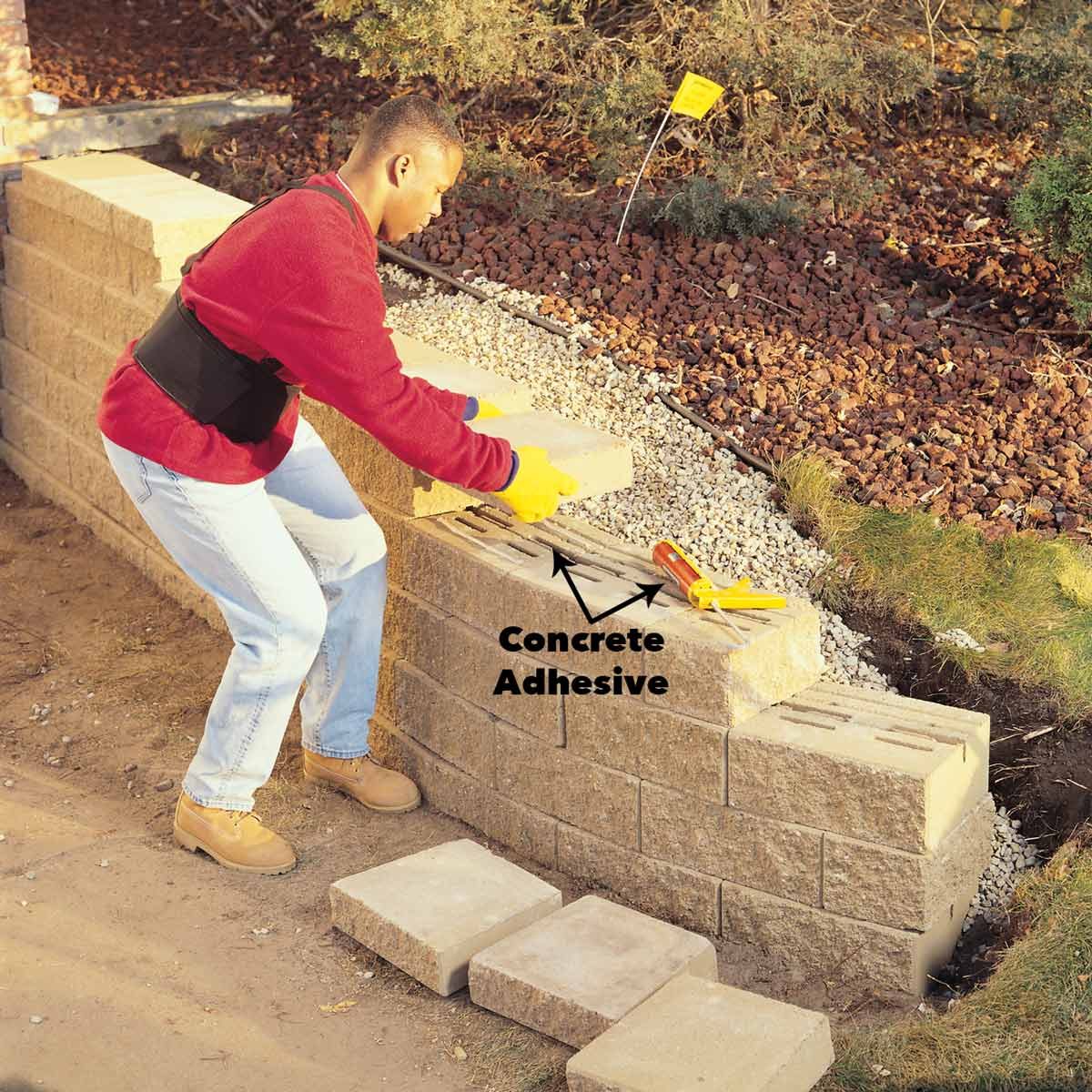 A person is placing a concrete block onto a wall, using adhesive, in a landscaped outdoor area with gravel and grass.