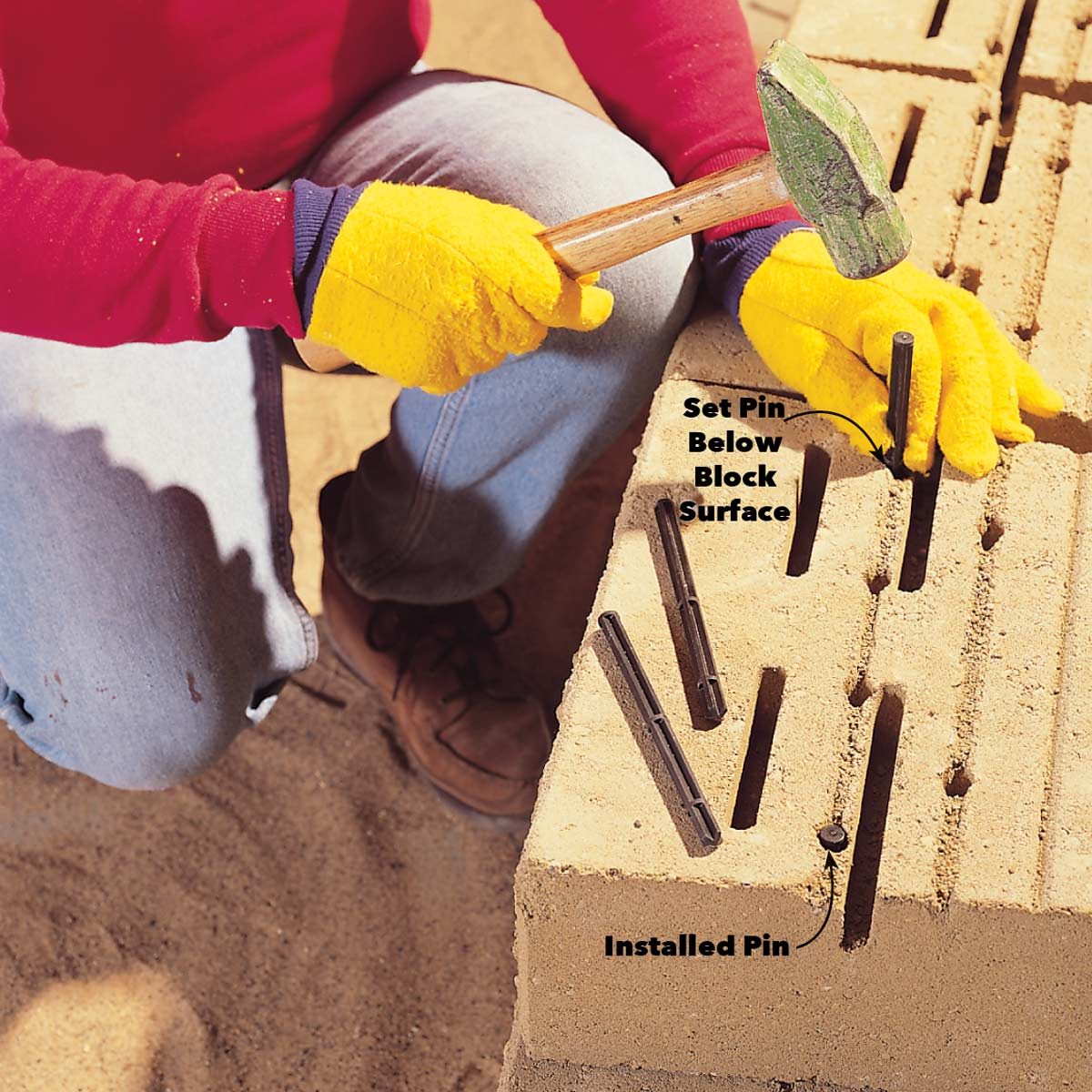 A person kneels, using a hammer to drive a pin into a concrete block, with several pins positioned nearby in a sandy environment.