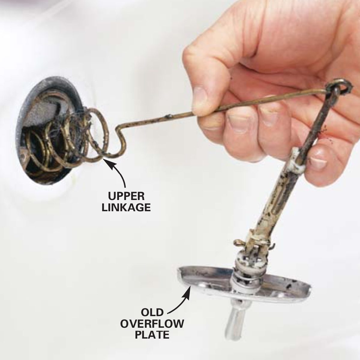 A hand holds an old overflow plate attached to linkage while removing it from a drain, set against a light-colored background.