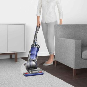 A vacuum cleaner is being pushed over carpet, while a person in light clothing stands beside a gray couch in a minimalistic room.