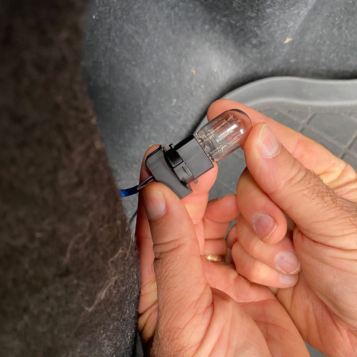 A person holds a light bulb socket with a transparent bulb, preparing to connect it in a dimly lit vehicle interior with a textured floor mat.