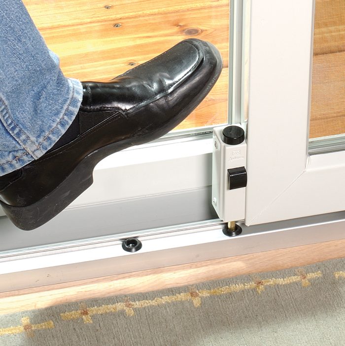 A black shoe presses down on a sliding door's locking mechanism, while wooden flooring and a patterned rug provide the surrounding context.