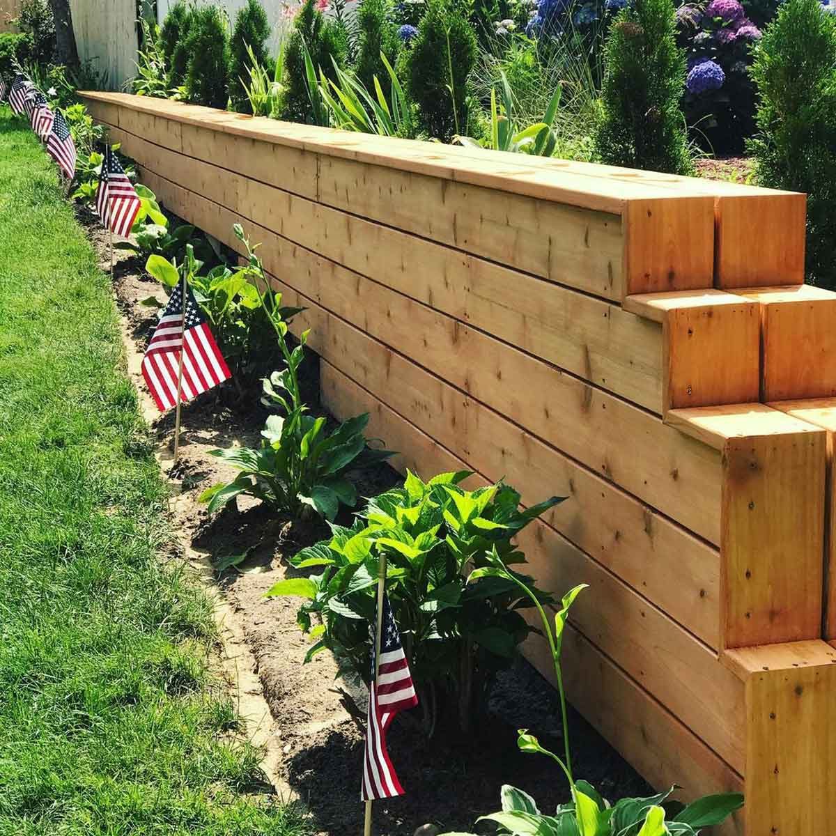 Wooden landscaping timbers form a long raised bed. Small American flags are placed along the planted area. Lush greenery surrounds the structure in a sunny yard.