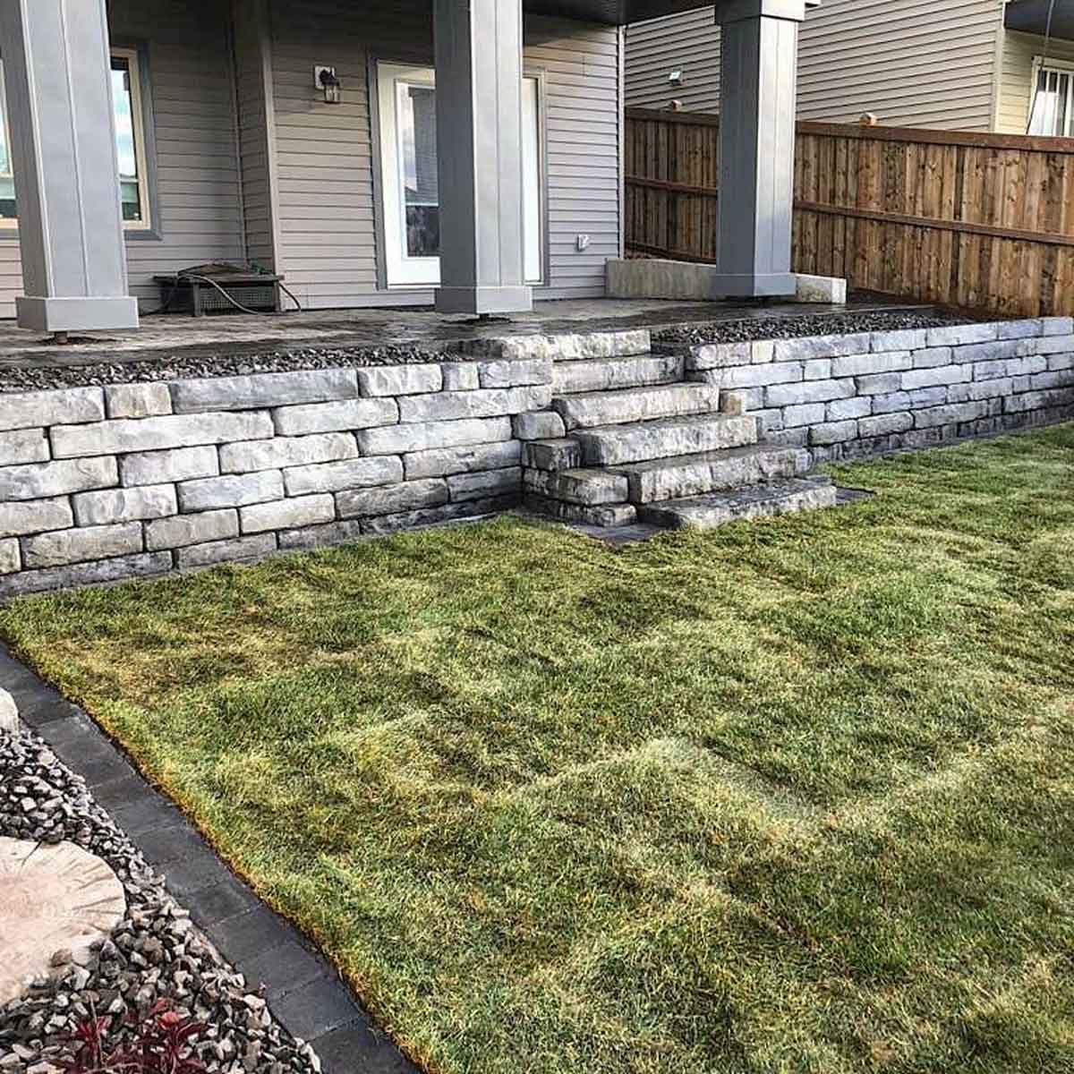 Stone steps lead from a raised patio to a grassy lawn, set against a backdrop of a wooden fence and house walls.
