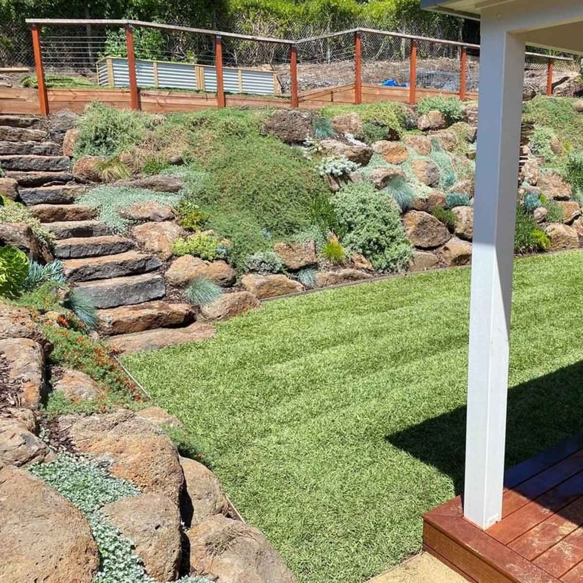 A landscaped garden features stone steps leading to a grassy area surrounded by lush greenery and rocks, under bright, sunny conditions.