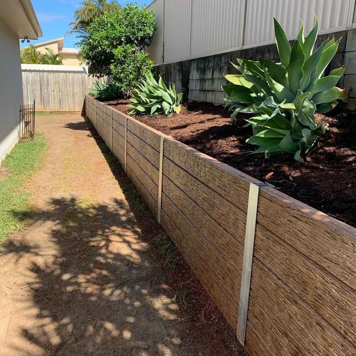 A wooden retaining wall lines a narrow dirt pathway, with green plants growing in a garden bed at the side, under a bright blue sky.
