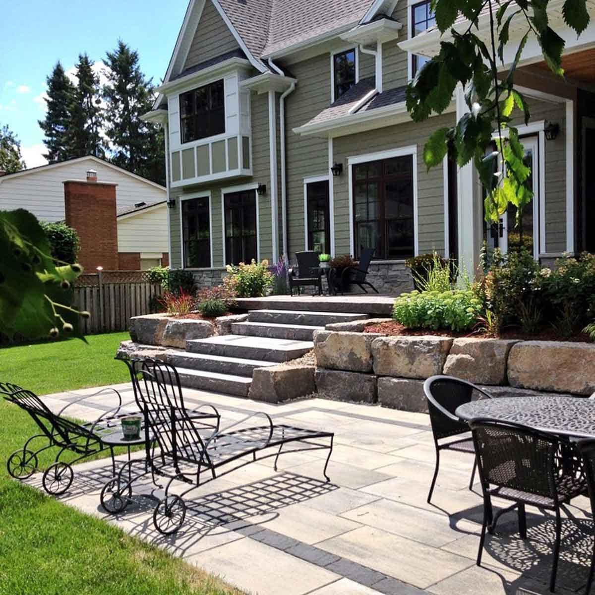 A patio with black metal chairs and tables features stone steps leading to a house surrounded by greenery and colorful plants under a clear blue sky.