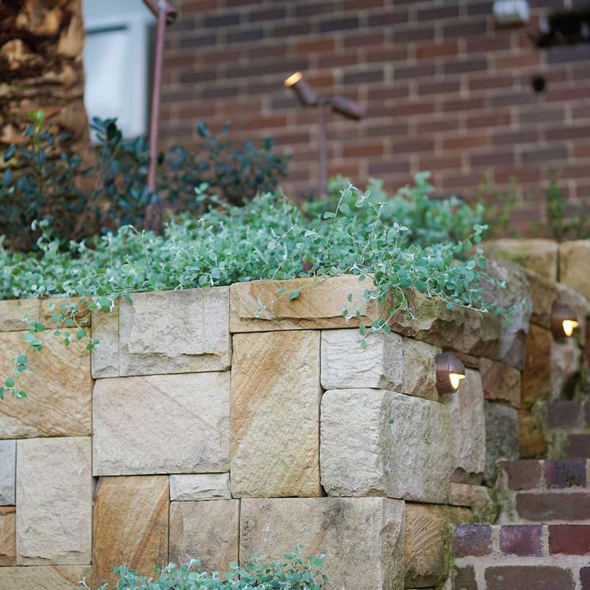 A stone planter holds lush green plants, while low lights illuminate its edge. A brick staircase leads upward, surrounded by a textured brick wall.