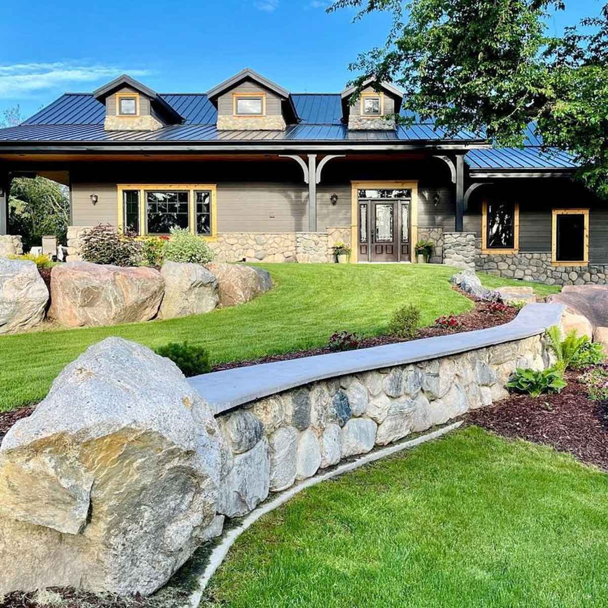 A stone house with a sloped roof sits amidst a manicured green lawn, featuring a curved stone wall and ornamental plants, under a clear blue sky.