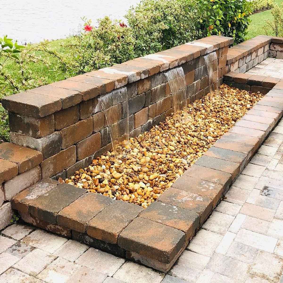 A stone water feature flows water over rocks, set in a landscaped area with greenery and pavers surrounding it, creating a tranquil outdoor space.