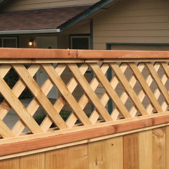 A wooden fence with a lattice top defines a boundary, positioned in front of a house with a roof and porch, surrounded by grass.