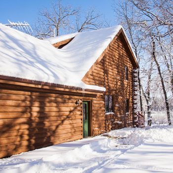 A wooden cabin is covered in snow, standing beside a snowy path, with leafless trees surrounding it under a clear blue sky.
