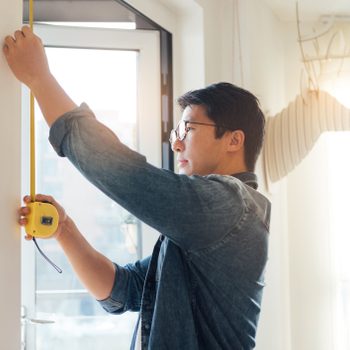 A man holds a tape measure against a wall, measuring height in a well-lit room with a window and a decorative antler-shaped object.