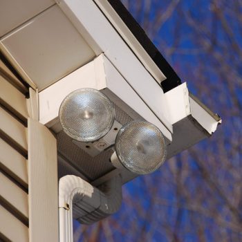 Two outdoor lights are mounted under a house's eave, shining downward, against a backdrop of a clear blue sky and bare tree branches.