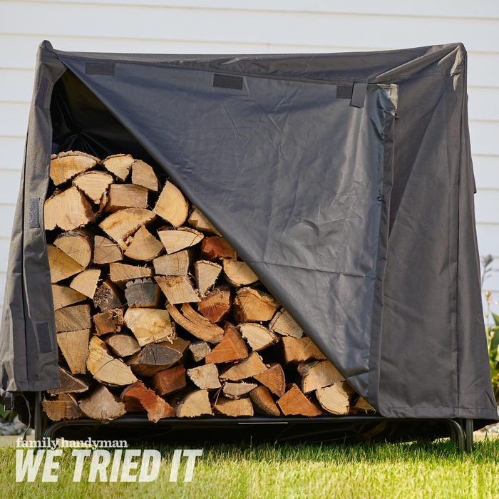 A covered woodpile displays neatly stacked logs, partially exposed under a black protective cover, set against a backdrop of grass and a light-colored building.