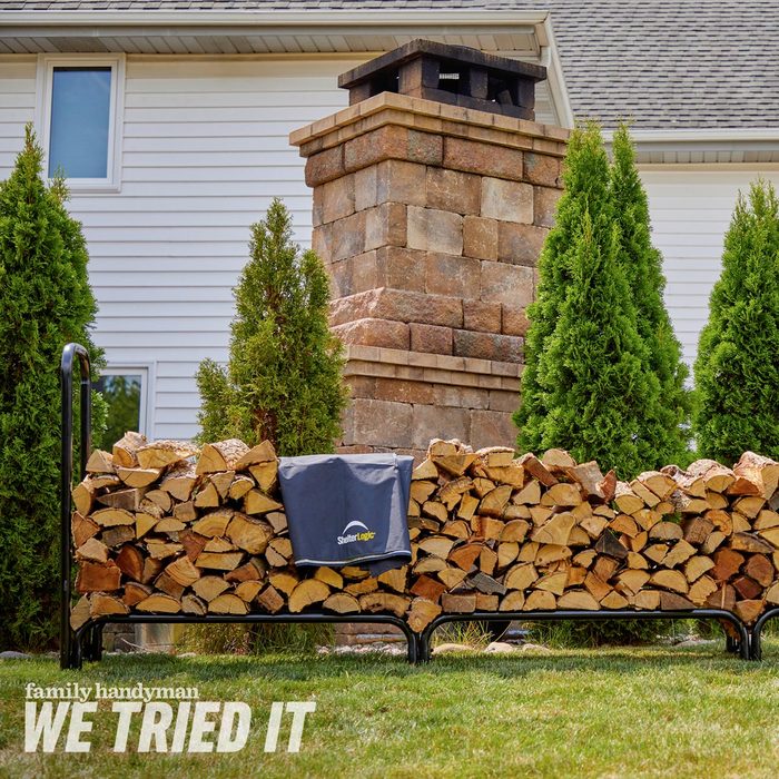 A wooden log rack holds neatly stacked firewood next to a stone chimney and green shrubs, set against a white house background under clear skies.