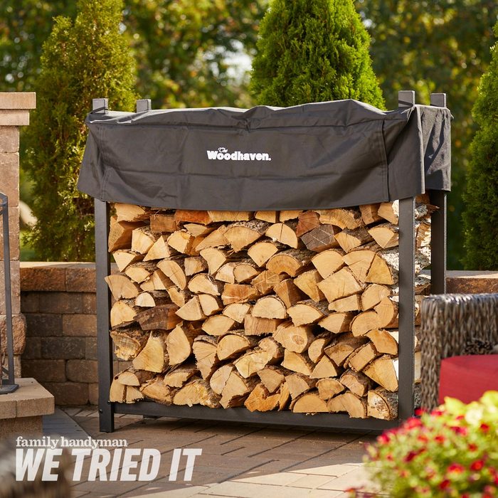 A wood storage rack holds neatly stacked firewood, covered with a black tarp. The setting includes greenery and a stone patio, creating an outdoor atmosphere.