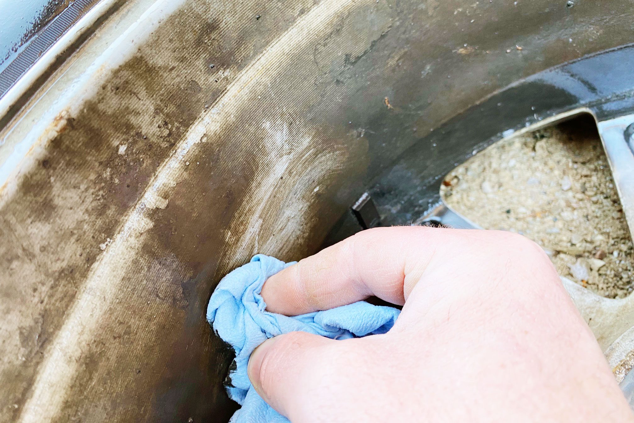A hand wipes a dirty metal surface with a blue cloth, revealing a cleaner area against a backdrop of grime and dirt.