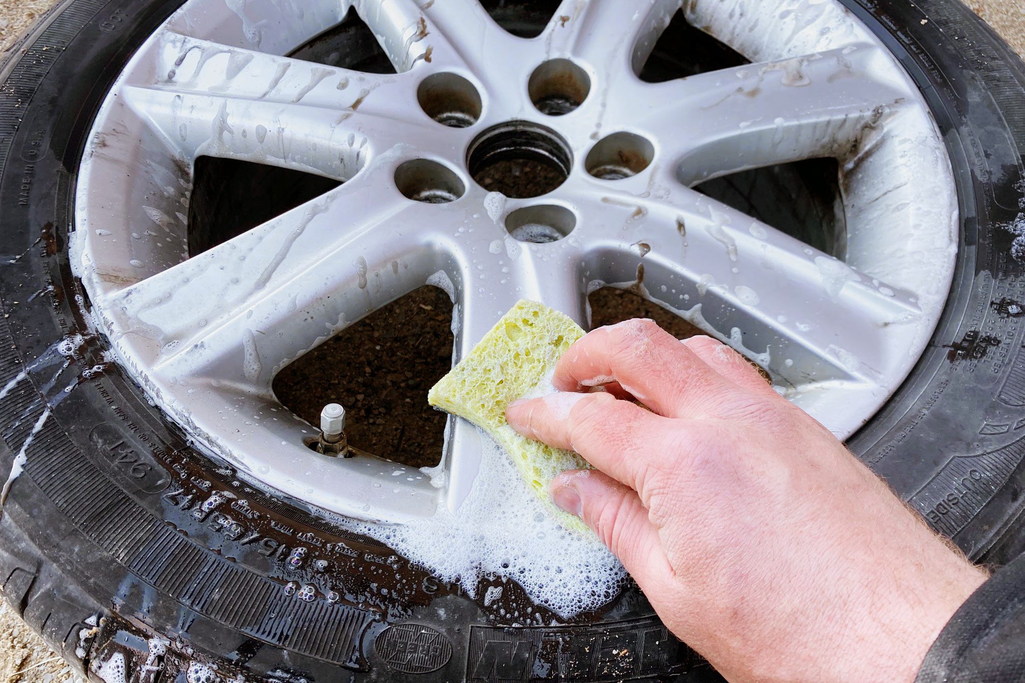 A hand scrubs a soapy, silver car wheel with a sponge, while dirt and bubbles accumulate on the tire's surface, indicating it's being cleaned.
