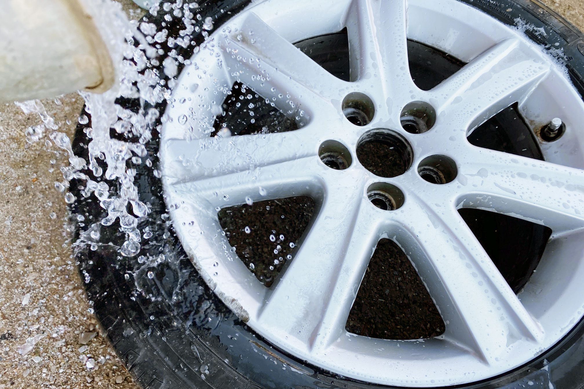 A silver alloy wheel is being rinsed with water, creating bubbles and droplets, while resting on a textured surface.